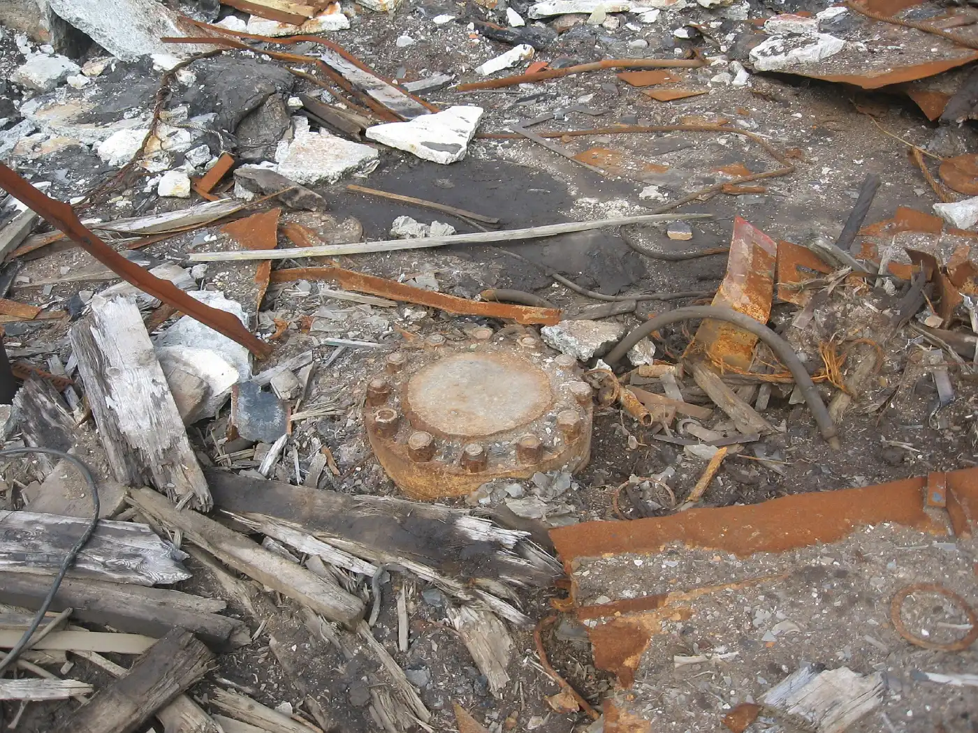 Close-up view of the rusted metal cover of the Kola Superdeep Borehole surrounded by scattered debris including rusted metal sheets, broken wood, rubble, and dirt.